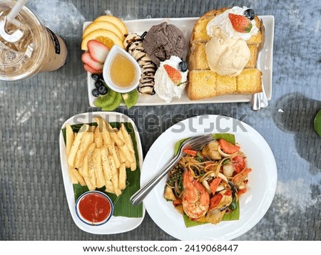 This picture shows a table with a variety of food on it. These foods include French fries, noodles, fruit, and ice cream. They are placed on plates and glasses. The table is located in the kitchen or 