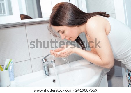 Beautiful woman in a white T-shirt washes her face with running water over the washbasin at home Royalty-Free Stock Photo #2419087569