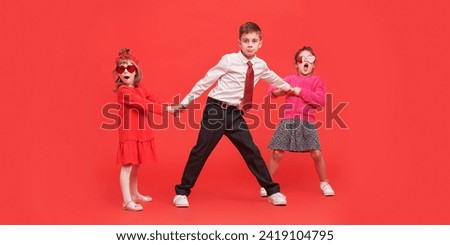 Two cute little girls in bright clothes and heart-shaped glasses and a boy in smart clothes pose emotionally on a red background. Full-length portrait. Kids fashion. 