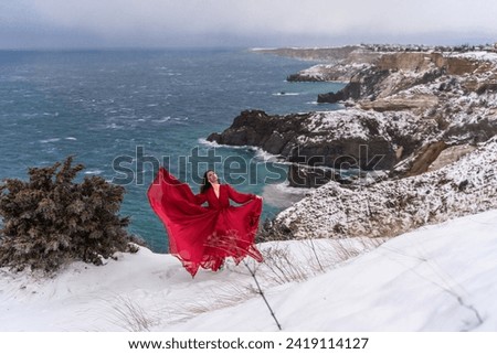 Woman red dress snow sea. Happy woman in a red dress in the snowy mountains by the emerald sea. The wind blows her clothes, posing against sea and snow background.
