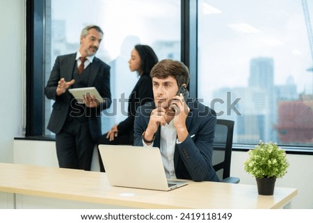 Focus at Young White businessperson using smartphone at desk with serious and thinking emotion while his co-worker talking at window in the modern office.