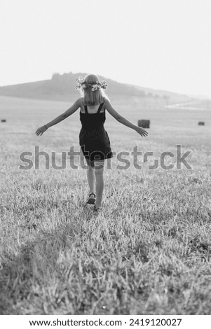 a girl with blond hair and a wreath of flowers on her head walks through the fields in summer, black and white photo