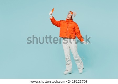 Full body young skier woman wear warm padded windbreaker jacket hat ski goggles mask do selfie shot on mobile cell phone travel rest spend winter season in mountains isolated on plain blue background