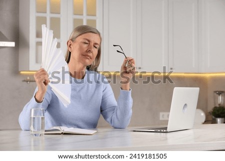 Menopause. Woman waving hand fan to cool herself during hot flash at table in kitchen Royalty-Free Stock Photo #2419181505