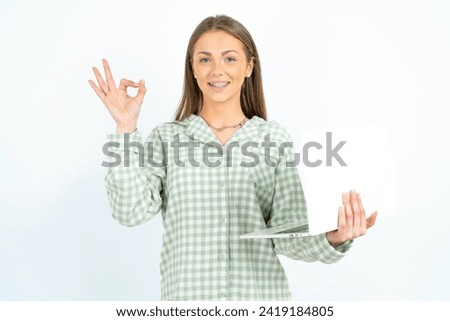 Positive Young beautiful woman wearing green plaid pyjama hold wireless netbook hand fingers show okey symbol