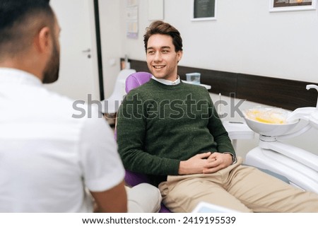 Portrait of cheerful bearded young man patient attentively listening unrecognizable male dentist discusses treatment plan sitting on dental chair in dentistry clinic. Concept of dentistry, teeth care. Royalty-Free Stock Photo #2419195539