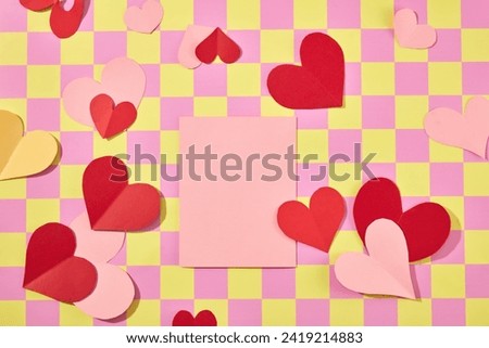 On pink and yellow checkered background, color paper cut in heart shape decorated with pink card in the middle. Top view, blank space for design. Valentine’s day concept