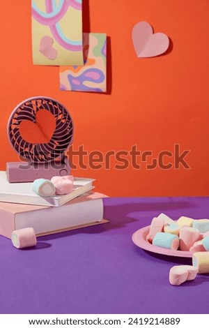 A stack of two books and an electronic fan placed on top featured. Pink dish containing lots of colorful marshmallows. Art pictures pinned on orange background