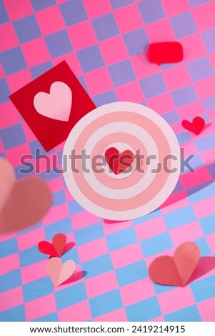 Front view of cute bullseye decorated with red paper hearts and card decorated on pink and purple checkered background. Minimal scene for advertising on valentine’s day
