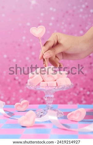 Front view of a glass tray full with pink marshmallow and ribbon decorated on pink background. A woman's hand is holding a candy stick and sticking it into the tray. Creative idea for advertising Royalty-Free Stock Photo #2419214927