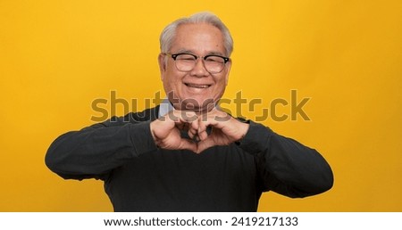 An old Asian man making a heart shape with his hands. Isolated on yellow background in the studio.