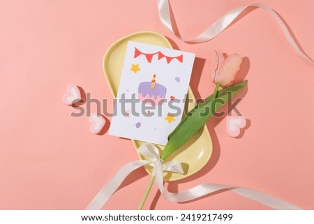 Close-up of a cute birthday card and a tulip on a yellow ceramic tray, next to a heart-shaped marshmallow and ribbon on a pink background. Birthday theme for advertising. Royalty-Free Stock Photo #2419217499