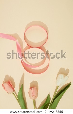 A ribbon in pink color arranged in a figure eight over beige background with few tulip flowers. Elegant card for International Women's Day. Banner or leaflet for March 8