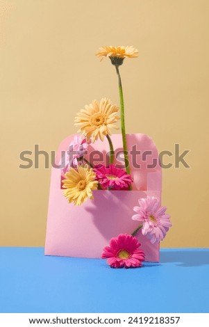 Brightly colored gerbera flowers are decorated around a pastel pink envelope on a minimalist background. Gerbera flowers mean fortune and luck.
