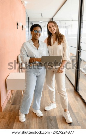 Two diverse female coworkers is using laptop, standing at the office. They holding laptop, looking at camera and smiling, full lengths, friendly office atmosphere and teamwork concept Royalty-Free Stock Photo #2419224877