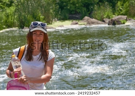 Young woman tourist with with a bottle of mineral water.Bank of a rapid river                                Royalty-Free Stock Photo #2419232457