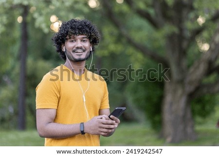 Portrait of a smiling Indian man doing sports and jogging in the park. Standing in headphones, holding a phone and smiling at the camera. Close-up photo.