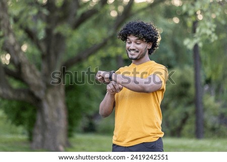 Photo of a smiling fashionable Indian man doing sports and running in the park. Stands and looks at the smart watch on his hand, checks the pulse, detects the time.