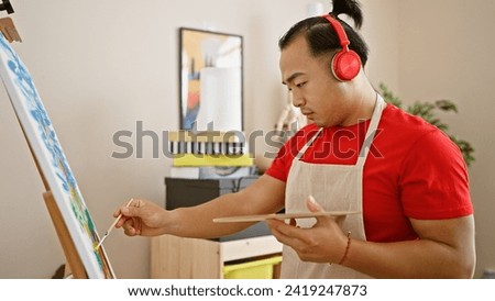 Serious young chinese man with pigtail hairstyle, artist standing in university art studio, effortlessly mastering the art of drawing while intently listening to music on headphones.