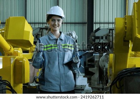 Caucasian female robotic engineer wearing helmet and safety glasses holding clipboard gives thumbs up showing awesomeness for industrial robot arm in factory.Innovation and technolgy for welding.