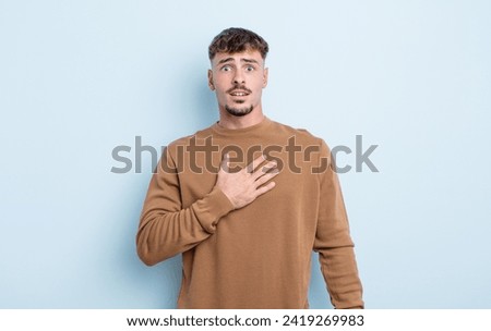 young handsome man feeling shocked, astonished and surprised, with hand on chest and open mouth, saying who, me? Royalty-Free Stock Photo #2419269983