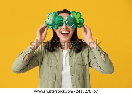 Beautiful young woman with decorative glasses in shape of clover on yellow background. St. Patrick's Day celebration