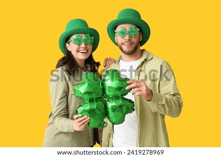 Beautiful young couple in leprechaun hats and decorative glasses with air balloon in shape of clover on yellow background. St. Patrick's Day celebration Royalty-Free Stock Photo #2419278969