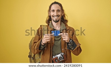 Young hispanic man tourist holding credit card smiling over isolated yellow background