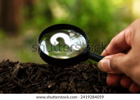 Hands holding magnifying glass focused in question mark sign on the soil ground in green garden. Concept of quiz, many question arising on soil ground with natural environmental background.