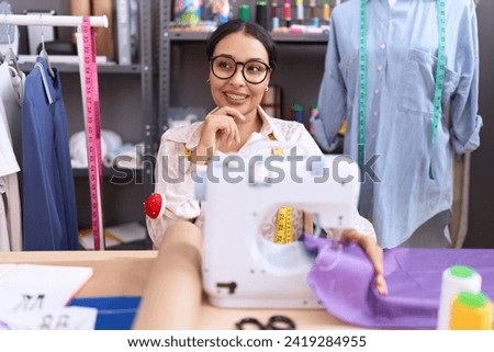 Young arab woman dressmaker designer working at atelier with hand on chin thinking about question, pensive expression. smiling and thoughtful face. doubt concept.  Royalty-Free Stock Photo #2419284955