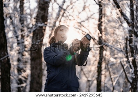 Young smiling woman photographer in outdoor gear looking up and taking photos on digital camera in winter snowy park, the girl takes pictures, outside photographer, travels