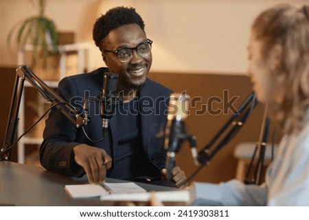 Medium shot of cheerful African American man wearing glasses and black suit talking to unrecognizable female guest sitting at table during podcast in studio