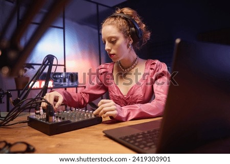 Medium shot of beautiful young woman with headphones switching channels on sound mixer during podcast recording