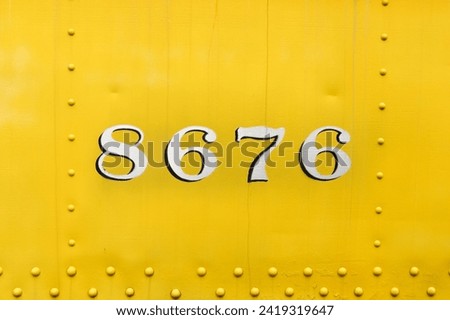 8676, stencil painted numbers on a vintage, antique train car. Yellow paint on thick sheet metal and four digits on a very old locomotive. Rivet rows surround the symbol