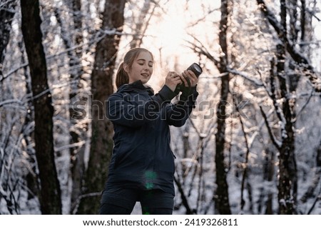 Young cheerful woman photographer in outdoor gear looking up and taking photos on digital camera in snowy park, outside photographer takes pictures in the winter forest, travels, holidays, hobby