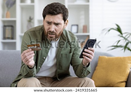 Upset cheated man sitting on couch in living room at home, holding bank credit card and phone, rejected money transfer, negative balance, scam money loss. Royalty-Free Stock Photo #2419331665