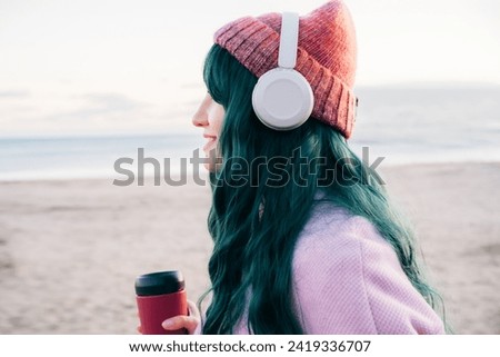 Side view young smiling hipster woman with color hair wearing wireless headphones, pink coat, hat, walking on beach with reusable coffee cup. Time To Relax. seasonal fashion style trend. Barbiecore Royalty-Free Stock Photo #2419336707