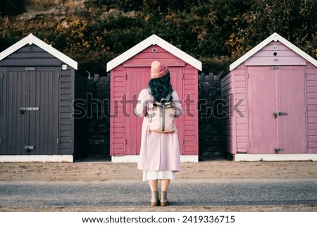 Back view stylish hipster woman with color hair in total pink outfit and backpack looking at wooden beach huts. Off season Travel concept. Seasonal street fashion. Barbiecore style. Simple pleasures Royalty-Free Stock Photo #2419336715