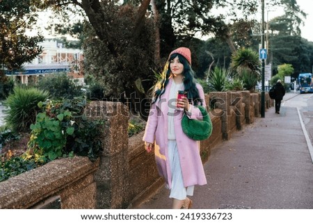 Stylish young smiling hipster woman with color hair walking on street in pink outfit with reusable coffee cup wearing coat, knitted hat, fur bag, happy mood, seasonal fashion, Barbiecore style trend Royalty-Free Stock Photo #2419336723