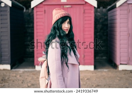 Stylish hipster woman with color hair in pink outfit and backpack walking along wooden beach huts on seaside. Off season Travel concept. Seasonal street fashion. Barbiecore style. Simple pleasures. Royalty-Free Stock Photo #2419336749