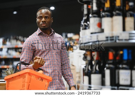 African American man holding bottle of wine and looking at it while standing in a wine store. Royalty-Free Stock Photo #2419367733