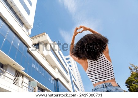 Rear view photo of funny cute lovely girl raise hands demonstrate heart shape love summer sunny weather big town outdoors