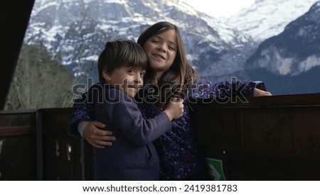 Young Siblings posing for photo standing in chalet balcony with mountains in background. Little brother and sister embrace during family vacation in the Alps