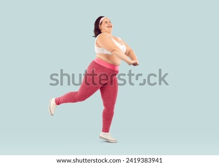 Full length shot of funny chubby overweight woman doing sports. Happy plus size, fat woman wearing sportswear doing physical exercises isolated on light blue studio background Royalty-Free Stock Photo #2419383941