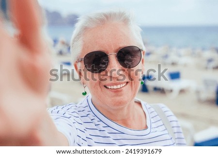 A senior woman with grey hair, in sunglasses taking a selfie on a beach 