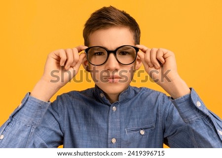 Focused teenage boy in denim shirt adjusting his large black glasses, nerdy teen male kid standing against mustard yellow background, embodying studious and intellectual vibe, closeup shot