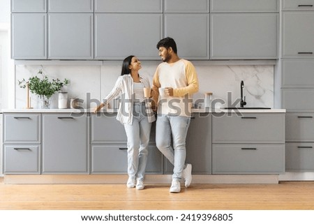 Cute cheerful loving young indian man and woman wearing homewear standing by table, drinking coffee together at kicthen in the morning, have conversation, smiling, full length, copy space Royalty-Free Stock Photo #2419396805