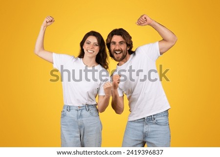 Enthusiastic european young couple in white t-shirts and jeans cheerfully raising fists in a victorious gesture, smiling with confidence against a bright yellow background