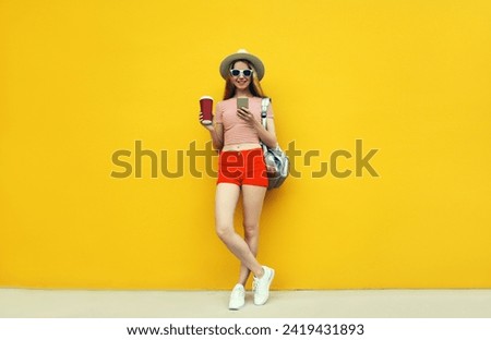 Summer image of traveler young woman 20s full length with mobile phone looking at device in casual straw hat with backpack on bright yellow background