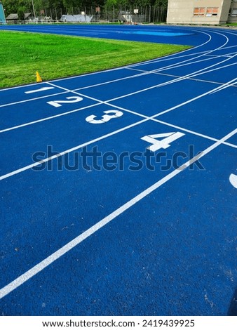 A blue running track with four lanes and numbers marking the starting line for a race. Green turf and a brick building with trees in the background. Isolated on a blue background. Royalty-Free Stock Photo #2419439925